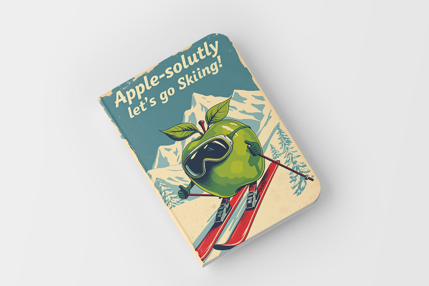 Notebook with a green apple wearing goggles and skis on a blue and beige cover.