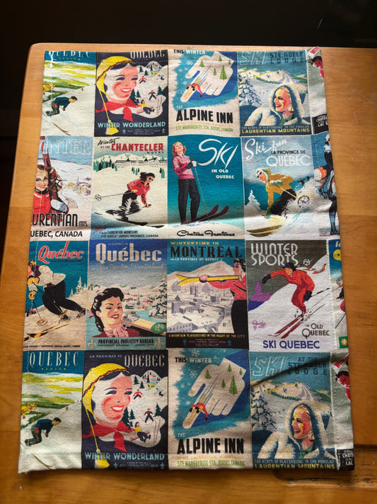 A towel  featuring a vintage collage print of Quebec ski resort posters. The print includes illustrations of skiers, snowboarders, and classic winter advertisements for places like Chantecler, Alpine Inn, and Winter Wonderland, displayed flat on a wooden surface.