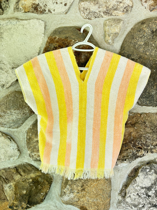A short-sleeve V-neck woven poncho or tunic with vertical stripes in yellow, peach, and off-white, hanging on a white plastic hanger against a rustic stone wall.